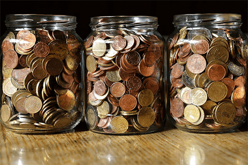 Saving money from buying in bulk-tycoonstory | tycoonstory media Three glass jars filled with assorted coins placed on a wooden surface, symbolizing savings, budgeting, and financial planning.