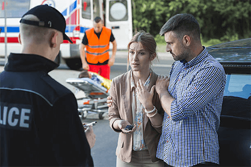 Car accident witness-tycoonstory | tycoonstory media Couple speaking with a police officer at the scene of a car accident, with an ambulance and emergency personnel in the background.