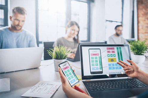 Tips to choose a mobile app development company-tycoonstory | tycoonstory media A team of developers working at an app development company, reviewing app interfaces on laptops and mobile devices to design user-friendly applications.
