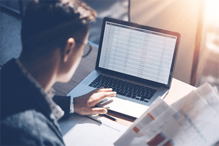 Accounting terms-tycoonstory | tycoonstory media Person wearing glasses looking at a laptop screen filled with data/spreadsheets while holding documents, representing someone working with financial data or reviewing accounting terms and records.