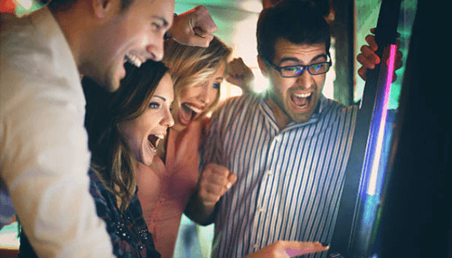 Analyze before you place your bets - tycoonstory | tycoonstory media Group of friends celebrating and cheering together while playing a slot machine, showing excitement and joy in a casino setting.