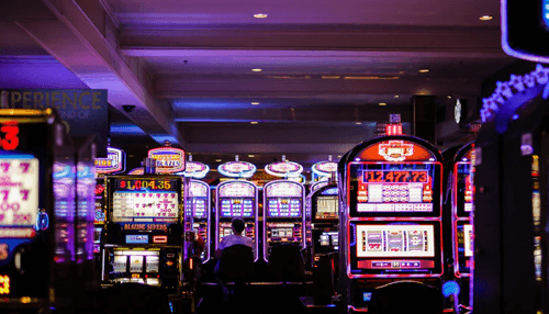 How to stream online casino games - tycoonstory | tycoonstory media A wide shot of a dimly lit casino floor with rows of brightly lit slot machines. A person is sitting in the center foreground, playing one of the slot machines under the neon and purple lights.