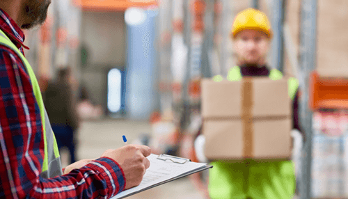 Returns labels with original packaging - tycoonstory | tycoonstory media Warehouse staff checking a clipboard as a worker carries returned boxes, illustrating operational processes involved in reverse logistics management.