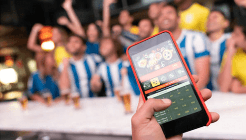 Gaming businesses - tycoonstory | tycoonstory media Smartphone displaying a betting app held in front of cheering sports fans, representing audience demographics and engagement in mobile betting.