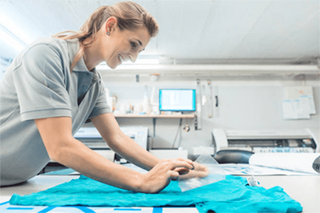 T-shirt printing methods A woman smiling while pressing a design onto a blue shirt in a workshop, showing a hands-on t-shirt printing business setup.