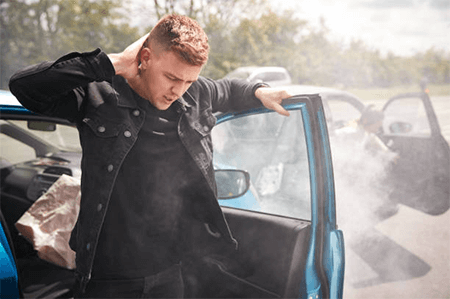 Steps to follow after a car accident-tycoonstory | tycoonstory media Man holding his neck while stepping out of a damaged car with smoke, showing signs of a car accident.