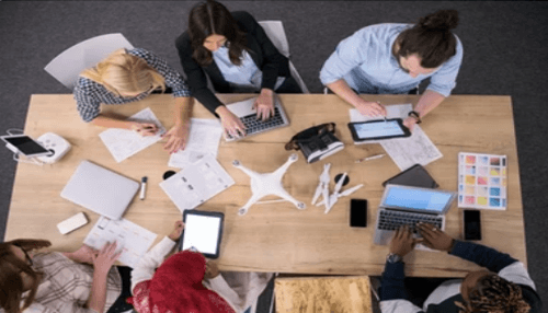 Create a business plan An overhead view of a team collaborating around a table covered in digital devices, documents, and drone hardware, representing the strategic planning phase of a professional drone service business.