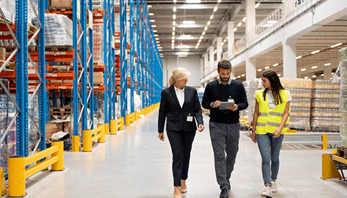 Warehousing technology processes Two managers and a worker in a safety vest discussing warehousing operations while walking through a bright aisle.