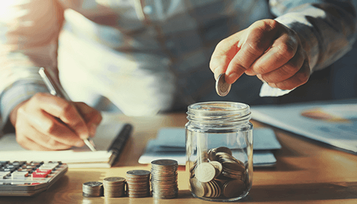 Outsource some tasks A person saving money by placing coins into a jar, illustrating the process of reducing overhead costs through smart financial planning.