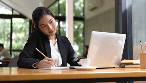 Qualified transcribers in your field A businesswoman taking notes while talking on the phone, symbolizing multitasking and effective transcription services for professionals.