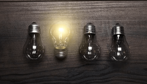 Uniqueness Four light bulbs placed in a row on a dark wooden surface, with one bulb glowing to represent a standout idea or innovation.
