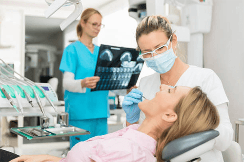Visit the dentist Dental professional examining a patient in a clinic while treating a broken or chipped tooth, with dental equipment and x-ray imaging visible in the background.