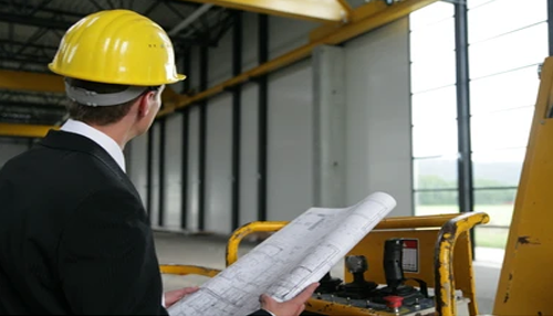 Building codes and safety regulations Engineer in a hard hat reviewing blueprints inside a large industrial facility during a business building construction project