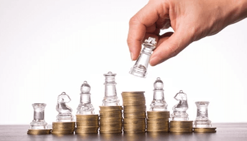 Investment decisions A hand strategically placing a clear glass chess king on top of a stack of gold coins, which are arranged in a bar graph formation alongside other chess pieces. This visual metaphor represents how strategic planning and investment rules can build and protect wealth.