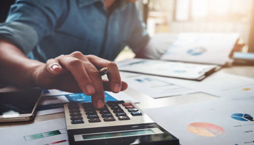 Cost analysis A close-up of a professional analyzing data with a calculator, documents, and a smartphone, demonstrating the precision required to maintain a balanced business budget.