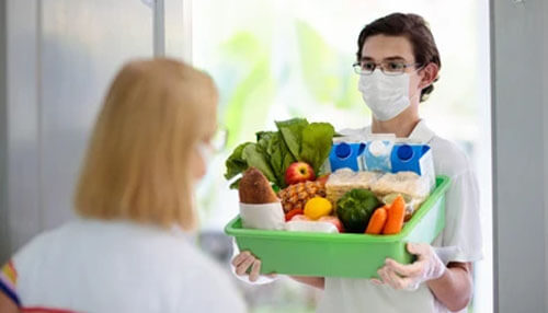 Grocery Delivery A delivery person with a face mask handing a box of groceries and essentials to a customer at the door.