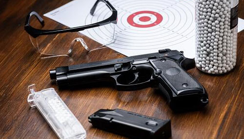Airsoft guns online Black airsoft guns handgun, safety glasses, a target, and a bottle of white plastic bb pellets on a wooden table.