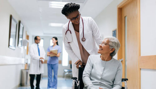 Nursing A compassionate healthcare provider in a white coat speaking warmly to an elderly patient in a wheelchair in a bright hospital hallway, showcasing patient-centered care in nursing.