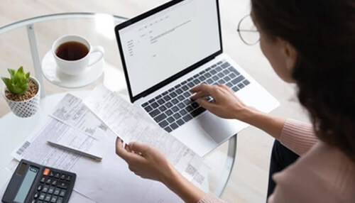 Calculate your expenses Self-employed individual calculating expenses by reviewing bills and receipts while working on a laptop at a home office desk.