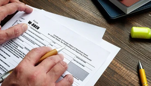 Role of the w-8ben form - tycoonstory | tycoonstory media Hands holding a pen over the w-8ben form for tax withholding. Focus on tax documentation.