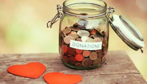 Donation jar filled with coins representing online fundraising for nonprofits, placed next to two red heart-shaped cutouts on a wooden surface Online fundraising for nonprofits