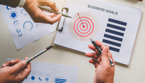 Break down each goal into specific steps that will help you reach it - tycoonstory | tycoonstory media A close-up, overhead view of three people's hands holding pens, pointing at a paper labeled "BUSINESS GOALS" which features a target graphic and a bar chart, demonstrating collaboration on strategic business goals.