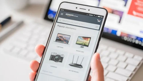 Competitor research Person browsing amazon on a smartphone to view popular electronics while working on a laptop in the background.