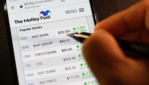 Motley fool A close-up of a mobile screen showing popular stocks from the motley fool website, with a person pointing to the stock details, indicating investment tracking.