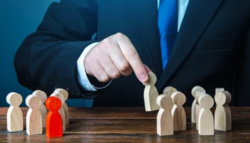 Recruitment sourcing strategies shown by a professional selecting a standout candidate from wooden figures.. Recruitment sourcing strategies