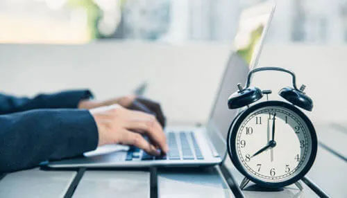 Person typing on a laptop with an alarm clock, representing time management and employee monitoring. Time tracking employee monitoring