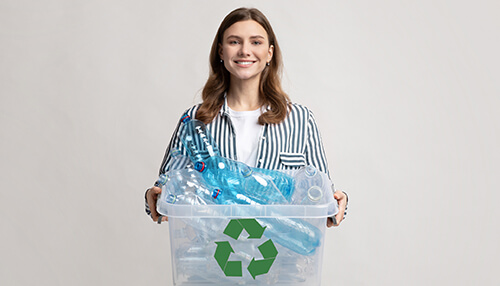Ban on plastic - tycoonstory | tycoonstory media Smiling woman holding a recycling bin filled with plastic bottles, to project eco-friendly waste management and recycling practices.