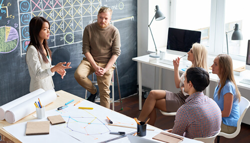 Team members having a collaborative discussion in a modern office setting with chalkboard diagrams and design materials on the table. Resource management