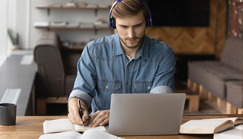 Virtual professional learning A man in a blue denim shirt and headphones focuses on a laptop screen, participating in a virtual professional learning session at a wooden desk surrounded by open reference books.