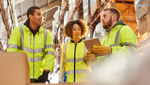 Warehousing costs - tycoonstory | tycoonstory media Business professionals discussing logistics in a warehouse setting, wearing safety gear and collaborating on work tasks, representing teamwork and efficiency in business operations.