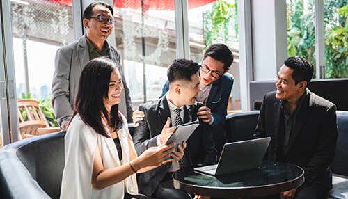 Always be willing to grow A group of five professionals in business attire gathered around a table, smiling and engaging in a discussion. One person is holding a tablet, while another is seated with a laptop. This collaborative environment is perfect for those aspiring to become a business tycoon.