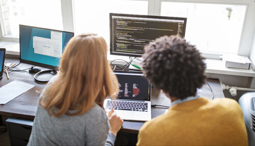 Field service management software - tycoonstory | tycoonstory media Two professionals facing each other in an office setting, reviewing and discussing field service management software on dual monitors.