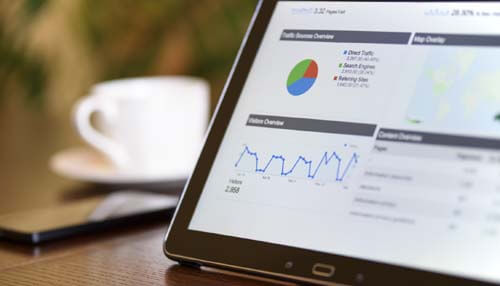 Become a business tycoon A tablet displaying a digital analytics dashboard with traffic and performance data, perfect for those looking to become a business tycoon. The background shows a cup of coffee and a smartphone, suggesting a productive work environment.