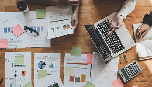 Pinpoint target customers Top-down view of a collaborative workspace with market analysis reports, a laptop, sticky notes, and office supplies.