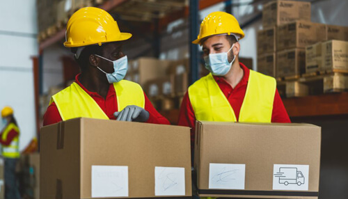 Reduction logistics operational cost - tycoonstory | tycoonstory media Two warehouse workers in reflective vests and helmets handling boxes signify efficient integrated logistics in product delivery.