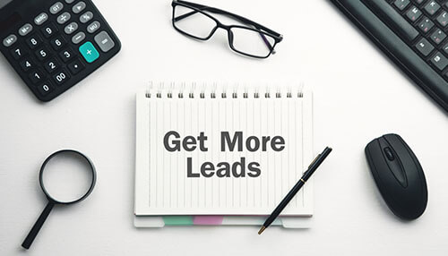 Create more leads - tycoonstory | tycoonstory media Notebook with the words get more leads placed on a desk along with a calculator, magnifying glass, glasses, pen, and mouse, representing a strategy for generating promotional products leads
