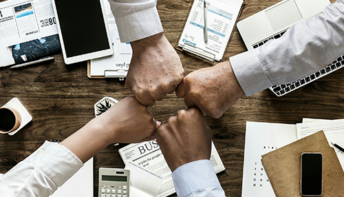 Make collaboration part of your culture Four people making a fist bump over a desk filled with business documents, laptops, and office supplies, symbolizing teamwork and collaboration in a business environment.