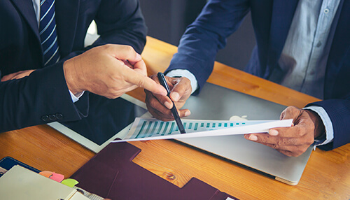 Purchase a domain for your business organization Two men in business suits discussing a document with charts over a wooden table, possibly reviewing metrics for an online store.