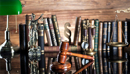 Legal trouble - tycoonstory | tycoonstory media A wooden gavel placed on a reflective surface with law books and symbols, representing legal compliance and the importance of rule of law in business.