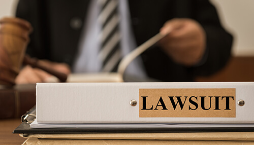 Lawsuits - tycoon | tycoonstory media Close-up of a binder labeled'Lawsuit' on a desk with a lawyer working in the background.