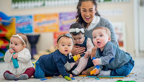 Responsibility of the daycare - tycoonstory | tycoonstory media Babies playing with toys on the floor while a caregiver smiles and supervises them at a cheerful daycare facility.