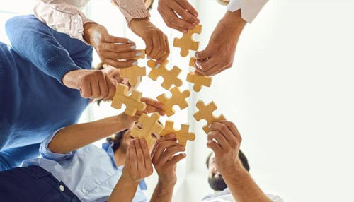 Solving a puzzle - tycoonsory | tycoonstory media Group of people holding wooden puzzle pieces together in the air, symbolizing teamwork and collaboration.