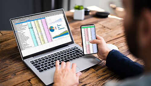 Using a spreadsheet - tycoonstory | tycoonstory media Business budget analysis on a laptop and smartphone displaying financial spreadsheets and graphs.