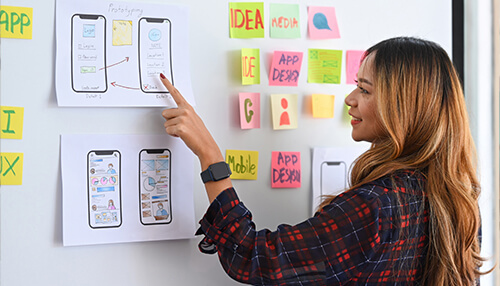 Develop Simple Apps only in the Manner of a Smartphone A user interface designer’s desk featuring a laptop, color palette swatches, and multiple hand-drawn mobile phone wireframes on paper, illustrating the strategic planning phase of Mobile App Development.