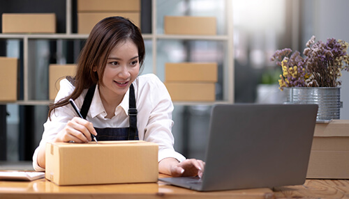 Identity what reselling business type to start A woman managing her online reselling business by preparing packages and working on a laptop in a well-organized workspace.
