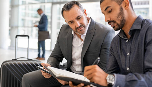 Inform your clients of your travel plans Business cheat sheet tip showing two professionals reviewing documents at an airport, highlighting client communication and travel planning strategies for business continuity.